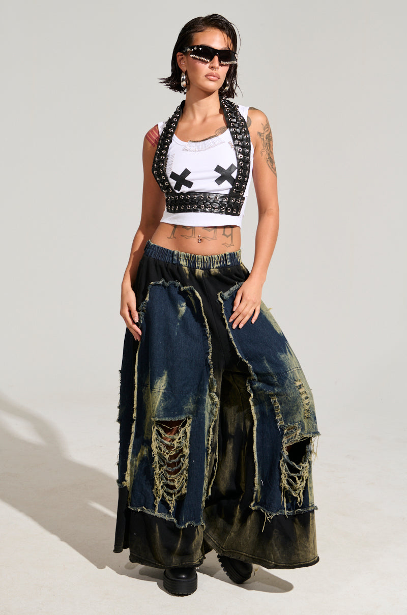 A person confidently showcases their edgy fashion style, wearing black sunglasses, a studded halter top, a white tank with black crosses, and the DOWN AND DIRTY DISTRESSED DENIM PANT featuring a wide-leg silhouette.