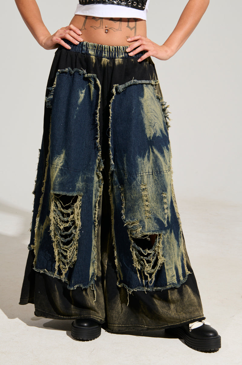 The individual is wearing the DOWN AND DIRTY DISTRESSED DENIM PANT, featuring a tie-dyed, wide-leg silhouette with large patches and frayed details. This unique style pant is paired with a cropped top and black platform shoes, revealing tattoos on their stomach against a plain background.