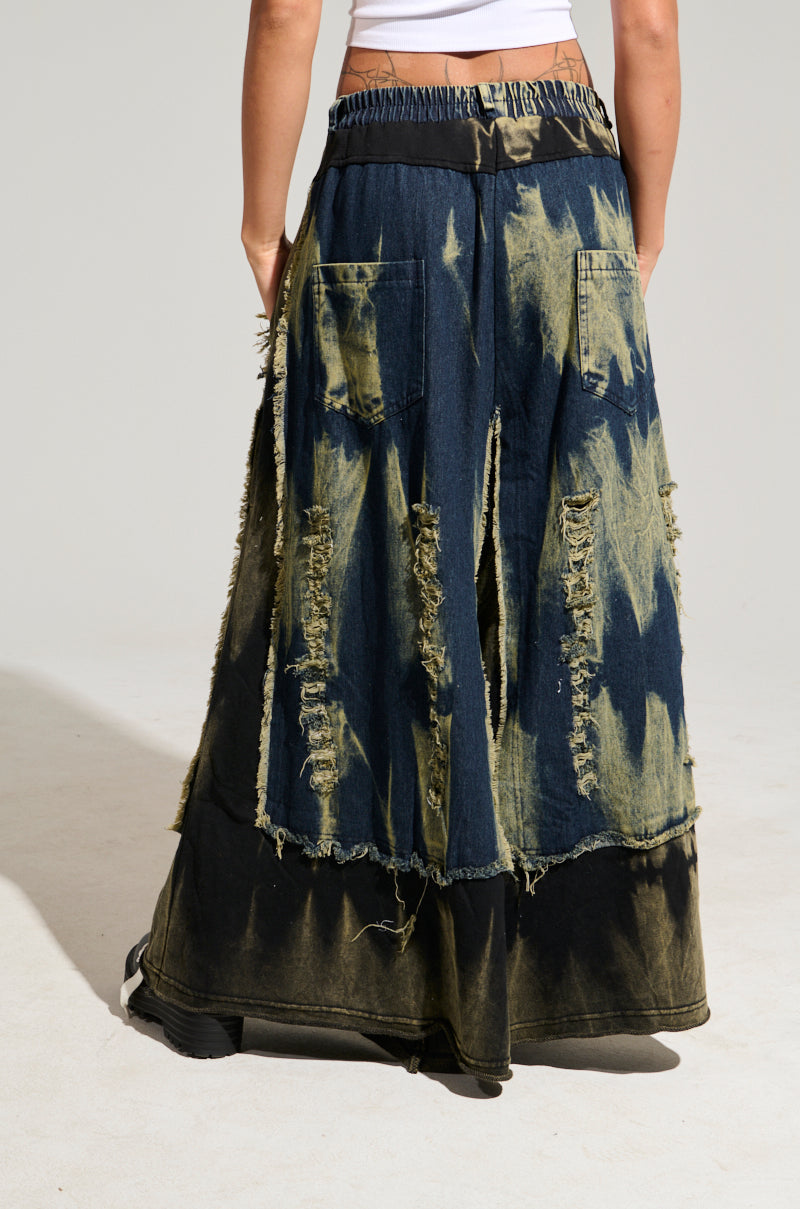 The DOWN AND DIRTY DISTRESSED DENIM PANT, featuring a wide-leg silhouette with large rear pockets and rugged edges in a tie-dye pattern of dark blue and beige tones, is worn by someone standing against a plain background.