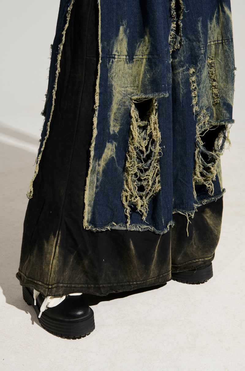 A close-up of the DOWN AND DIRTY DISTRESSED DENIM PANT showcases its wide-leg, dark denim design with an acid wash finish, embellished with frayed patches and asymmetrical hems. The wearer is standing in black platform shoes with white laces on a light surface.