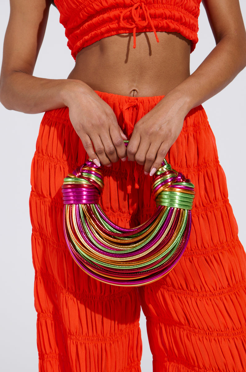 A person in a bright orange two-piece outfit, featuring a ruched crop top and pants with metallic detailing, holds the COAST TO COAST ROPE PURSE adorned with coiled, rainbow-colored bands against a plain white background.