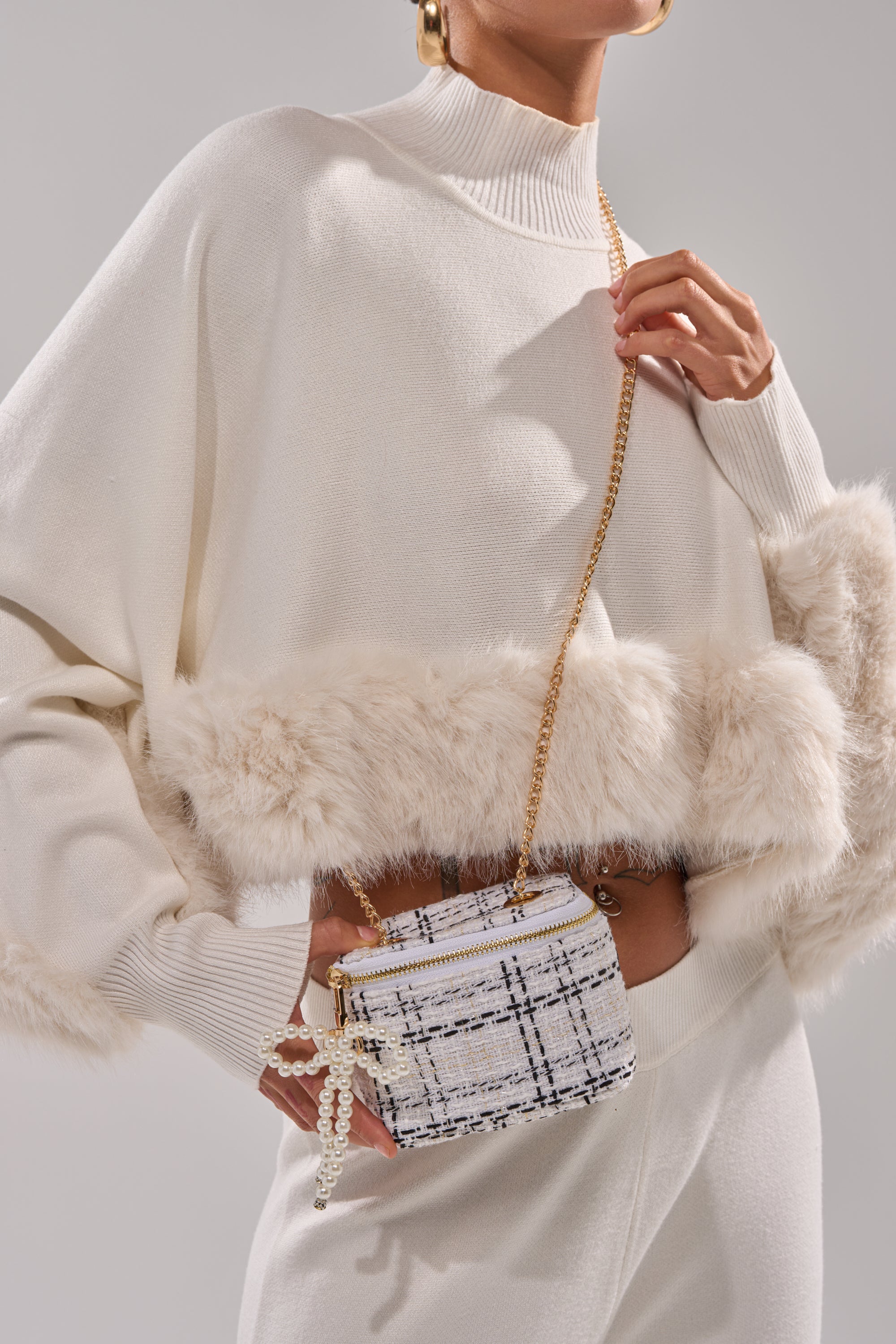 A person in a white, fur-trimmed sweater and matching pants holds the PEARLICIOUS MINI BAG, featuring pearl embellishments and a chain link strap, creating a chic look against a neutral background.