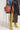 A person in a stylish outfit is holding the COURTSIDE BLING BASKETBALL PURSE encased in knotted rope. They are wearing a sheer black lace dress with a thigh-high slit, a denim jacket, and yellow snakeskin knee-high boots with stiletto heels.