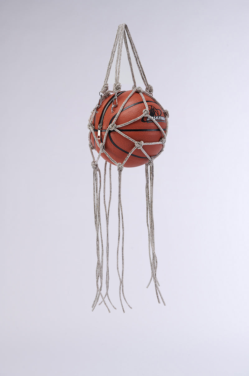 The COURTSIDE BLING BASKETBALL PURSE is intricately encased in a white macramé net, suspended in mid-air like an accessory fit for the court. The macramé net features multiple knots and hangs straight down, with several loose rope ends extending below the ball. The background is a plain, light grey.
