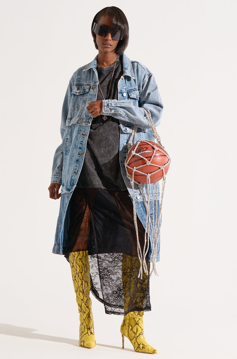 A person poses against a plain background wearing oversized black sunglasses, a long denim jacket, a dark sheer dress with lace detailing, and yellow snake print thigh-high boots. They are holding the eye-catching COURTSIDE BLING BASKETBALL PURSE, styled as a netted red ball.