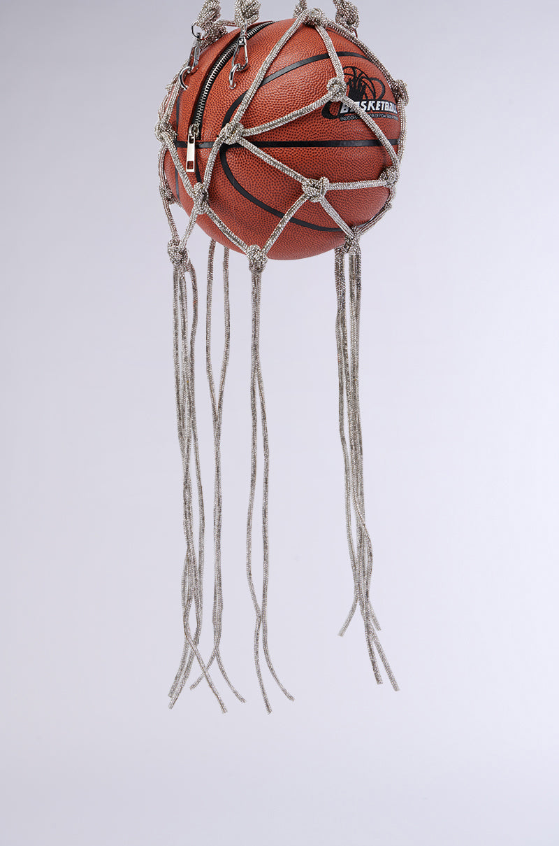 A basketball is suspended mid-air, stylishly entangled in a decorative macramé net. The net, reminiscent of the trendy COURTSIDE BLING BASKETBALL PURSE, is made of intricately knotted ropes with long, loose ends hanging downwards. The background is a plain, light grey color.