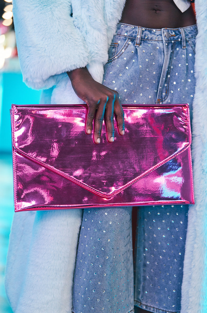 A person in a light blue fur coat and studded denim jeans holds a statement accessory—the NIKKI CHROME OVERSIZED CLUTCH in shiny pink. The background is blurred with blue and pink hues, emphasizing the bold choice of faux patent leather.