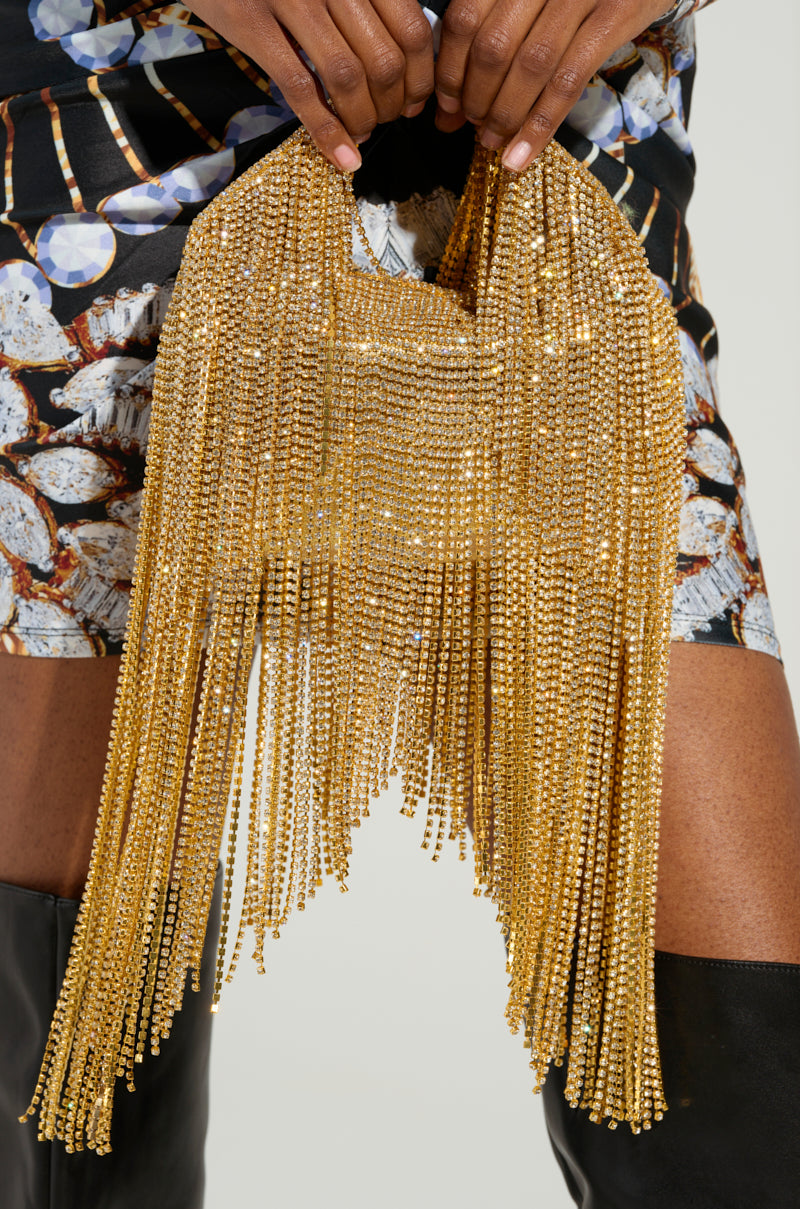 A person holding an ULTIMATE LUXE BLING PURSE, a luxurious gold accessory adorned with intricate beading and rhinestone fringe. They are dressed in a black and white patterned dress paired with tall black boots, set against a neutral background that accentuates the bag's shimmering details.