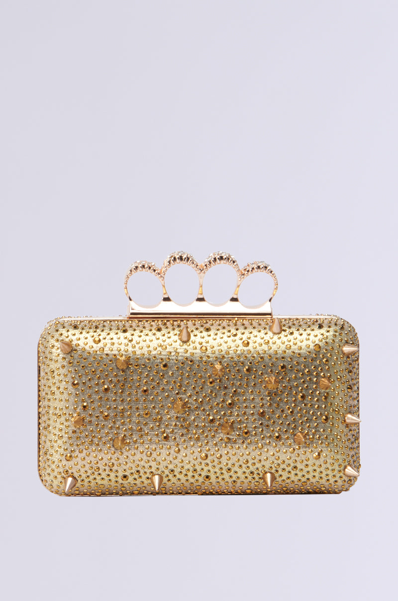Introducing the ALEXANDERS FAVE CLUTCH: a rectangular clutch in metallic gold, adorned with small studs and rhinestone-encrusted details. It features a distinctive four-ring handle embellished with intricate designs, offering a stylish and luxurious look.