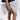 Standing against a plain white background, someone in a black outfit and lace-edged socks holds the GET WITH IT BOW CLUTCH. The clutch is adorned with a black and white polka dot print and features a metallic zip closure.