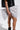 Standing against a plain white background, someone in a black outfit and lace-edged socks holds the GET WITH IT BOW CLUTCH. The clutch is adorned with a black and white polka dot print and features a metallic zip closure.