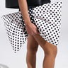 Standing against a plain white background, someone in a black outfit and lace-edged socks holds the GET WITH IT BOW CLUTCH. The clutch is adorned with a black and white polka dot print and features a metallic zip closure.