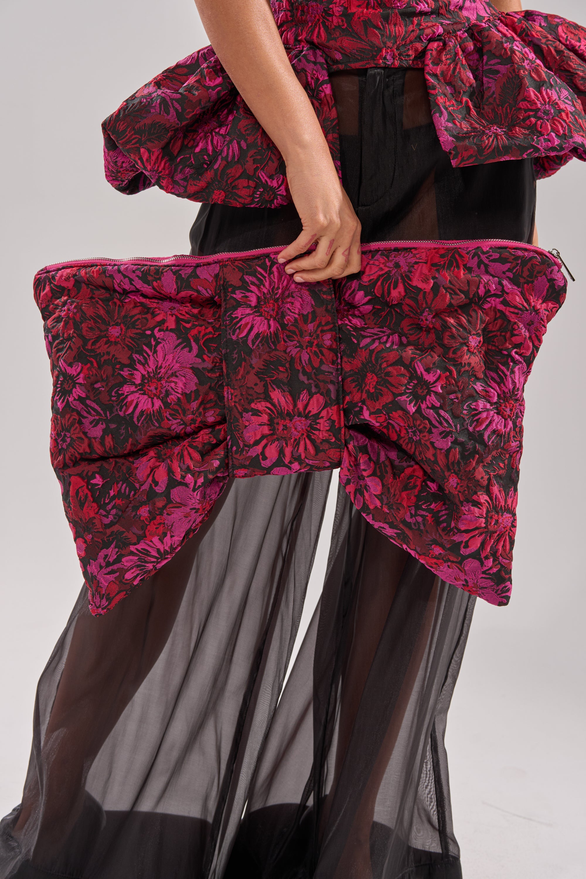 A person in sheer black pants holds the BOWTIFUL OVERSIZED CLUTCH, a bow-shaped purse with a red and pink floral pattern matching their top. The background is plain and light-colored. 3