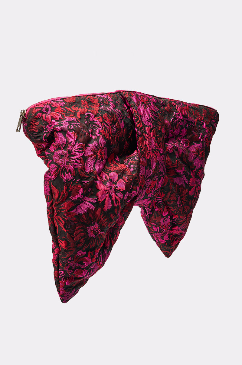The BOWTIFUL OVERSIZED CLUTCH is a bow-shaped handbag with a metallic zipper, featuring vibrant red and pink floral jacquard on a plain white background. 4