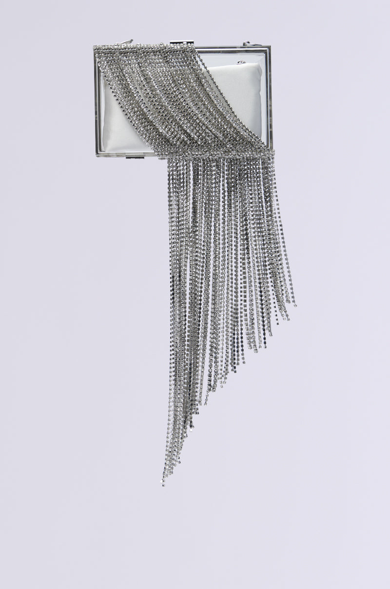 The OVER THA TOP CLEAR BLING CLUTCH features a rectangular silver design adorned with a shimmering diagonal rhinestone fringe on the front, set against a simple white background.