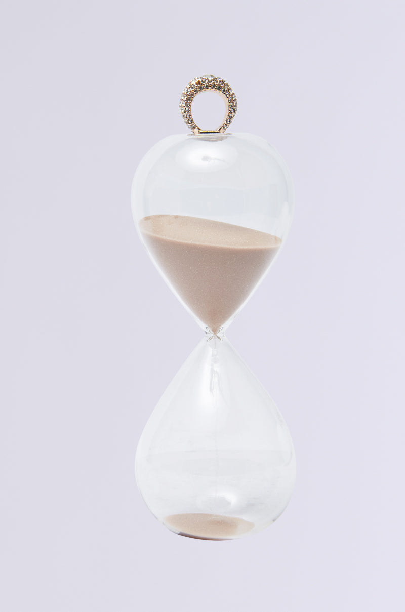 The WE GOT TIME SAND HOURGLASS RING showcases a transparent design with sand halfway through, flowing gently from the top half to the bottom. It features a decorative faux gold loop on top, set against a light gray background. 0