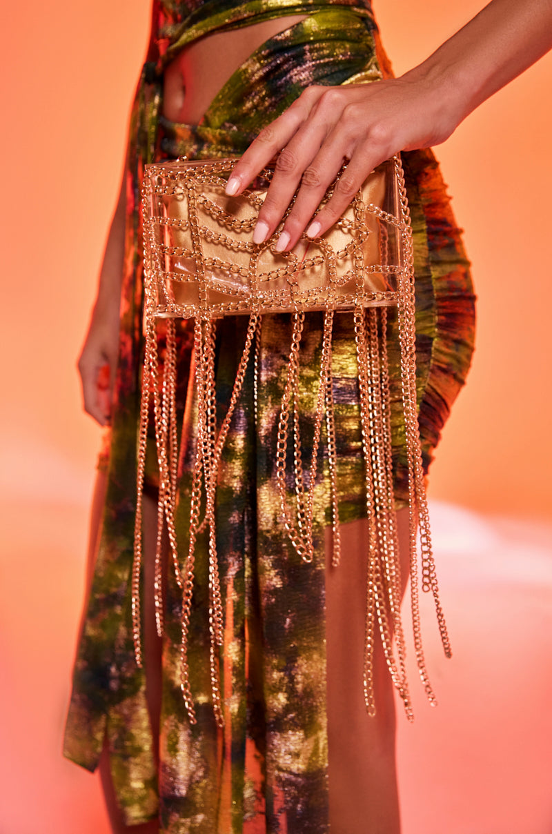 A person holding THE REAL RELATE CHAIN CLUTCH—a chic gold chain clutch with dangling chains—stands against an orange background, wearing a colorful ruched dress.