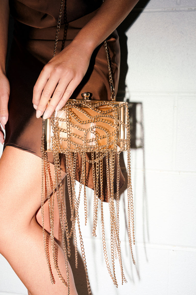 A person in a brown satin dress holds THE REAL RELATE CHAIN CLUTCH, a shiny gold purse with gold leather details and decorative gold chains, standing against a white textured wall.