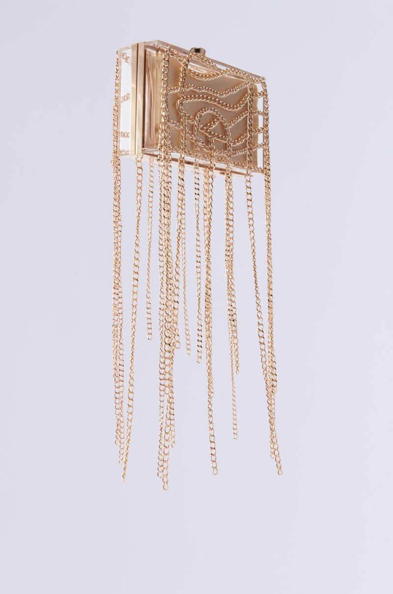 The REAL RELATE CHAIN CLUTCH is a rectangular, metallic gold piece with intricate patterns on its front and adorned with long dangling gold chains, resembling an acrylic hardshell, all set against a plain white background.