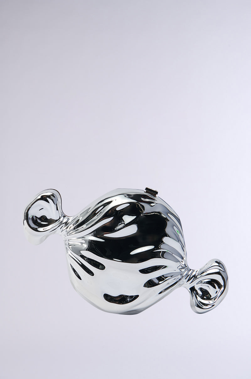 The TASTE LIKE CANDY CLUTCH, featuring a smooth and symmetrical design with twisted ends reminiscent of a classic wrapped sweet, sits centered against a plain white background. This metallic piece shines with a rhinestone-embellished surface that resembles wrapped candy.
