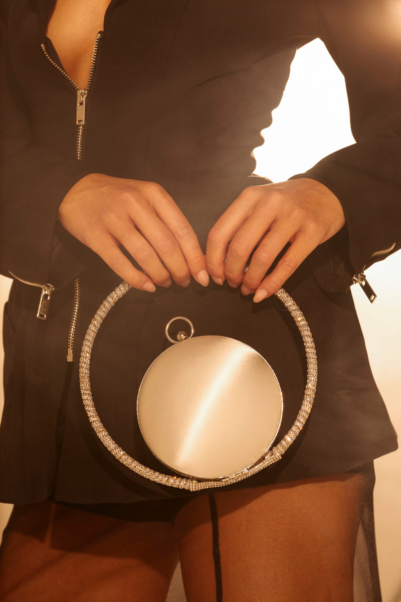 A person in a black zippered outfit holds the ITS MY WORLD PURSE, a spherical bag with a sparkling rhinestone handle, while standing in warm golden light.