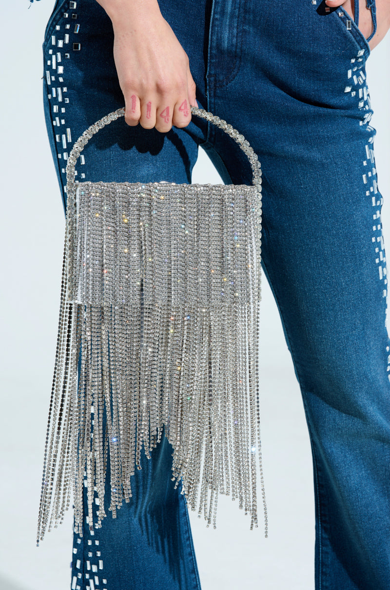 A person holds an EXPENSIVE TASTE BLING PURSE beside their studded denim jeans. The silver handbag features a rhinestone-adorned round handle and a long chainlink fringe trim cascading elegantly.