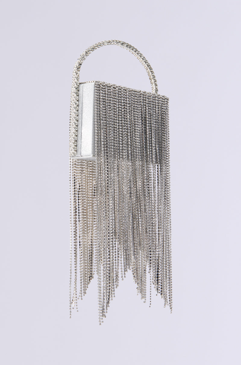 A rectangular silver handbag with dangling chains for a fringe effect and a handle dazzling with rhinestones exudes luxury. Perfectly complementing its name, the EXPENSIVE TASTE BLING PURSE stands out beautifully against a simple light grey backdrop.