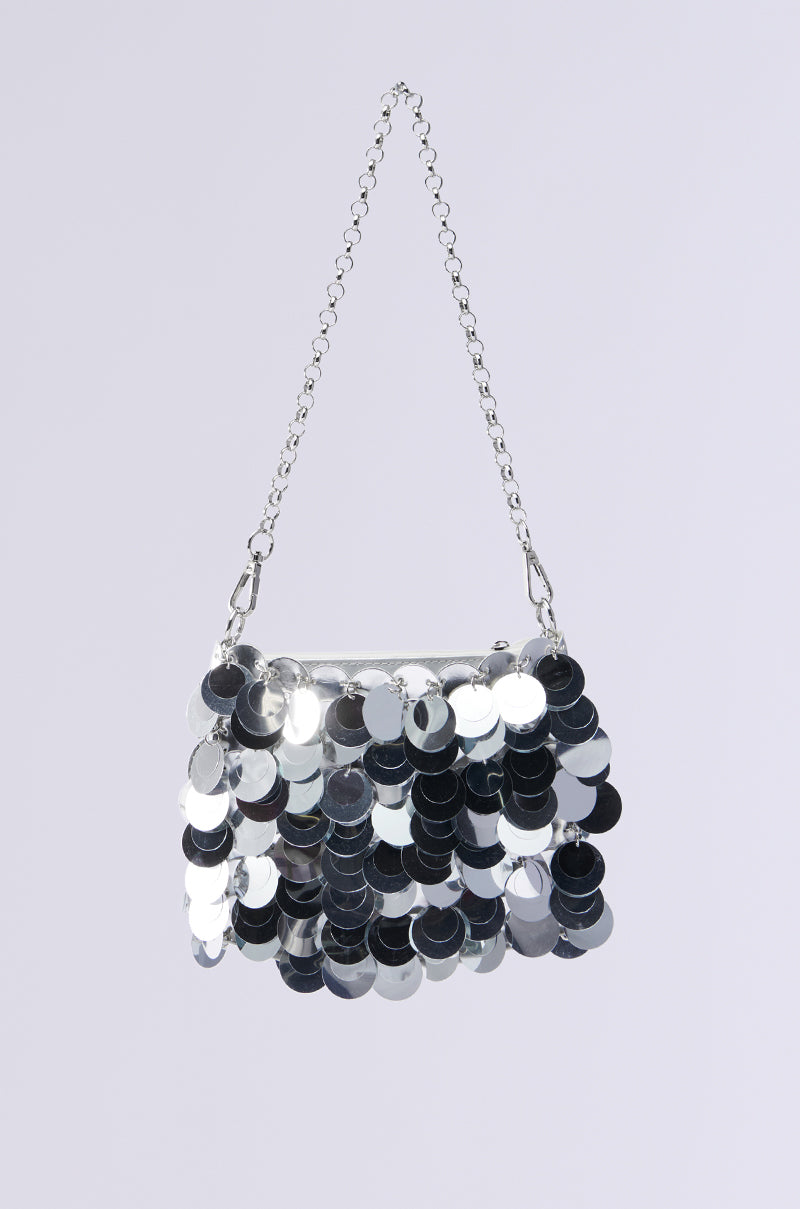 The DANCING THE NIGHT AWAY SEQUIN PURSE, featuring shiny silver sequins that overlap like fish scales for a reflective look, hangs elegantly from a delicate silver chain strap against a light gray background. This purse is the perfect accessory for any party.