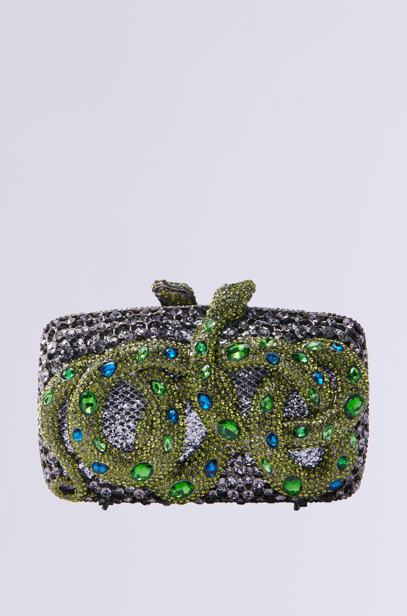 The "IN THE GARDEN RHINESTONE CLUTCH" is a rectangular bejeweled handbag with a metallic hardshell featuring an intricate snake design. Adorned with green, blue, and silver gemstones, the snake's sparkling green gems add a luxurious touch. 0