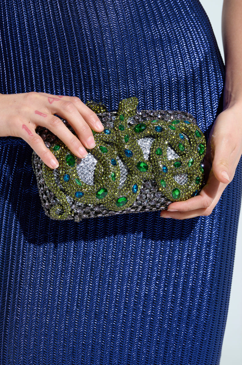 A person in a textured blue dress holds the IN THE GARDEN RHINESTONE CLUTCH, a glittering accessory adorned with green and blue gemstones forming a snake. 1