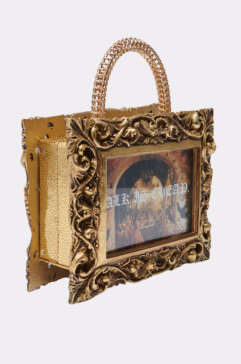 The TALK IS CHEAP FRAME BAG is a baroque-style handbag, resembling an ornate gold picture frame with detailed accents, a chain handle, a classical painting on the front, and "ALKSCHEAP" in white text.