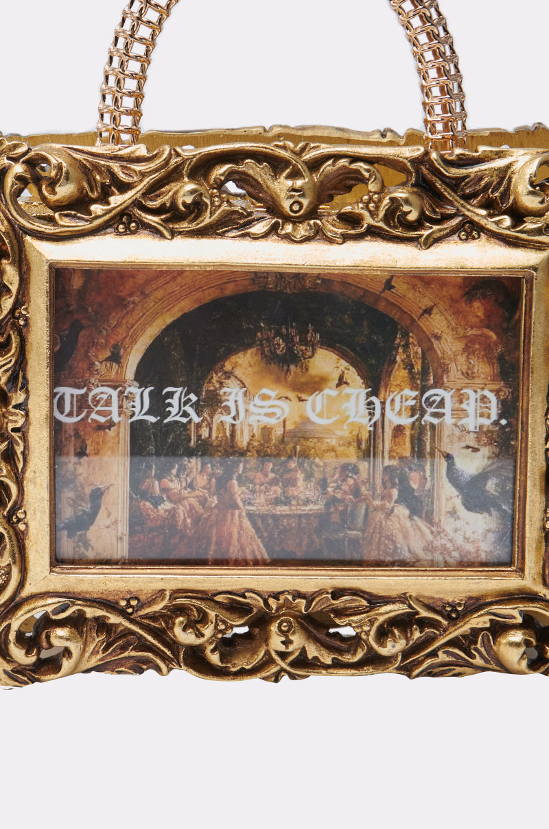 The TALK IS CHEAP FRAME BAG is a gold metallic bag shaped like a picture frame, with a vintage painting and "TALK IS CHEAP" in bold white letters across the center.