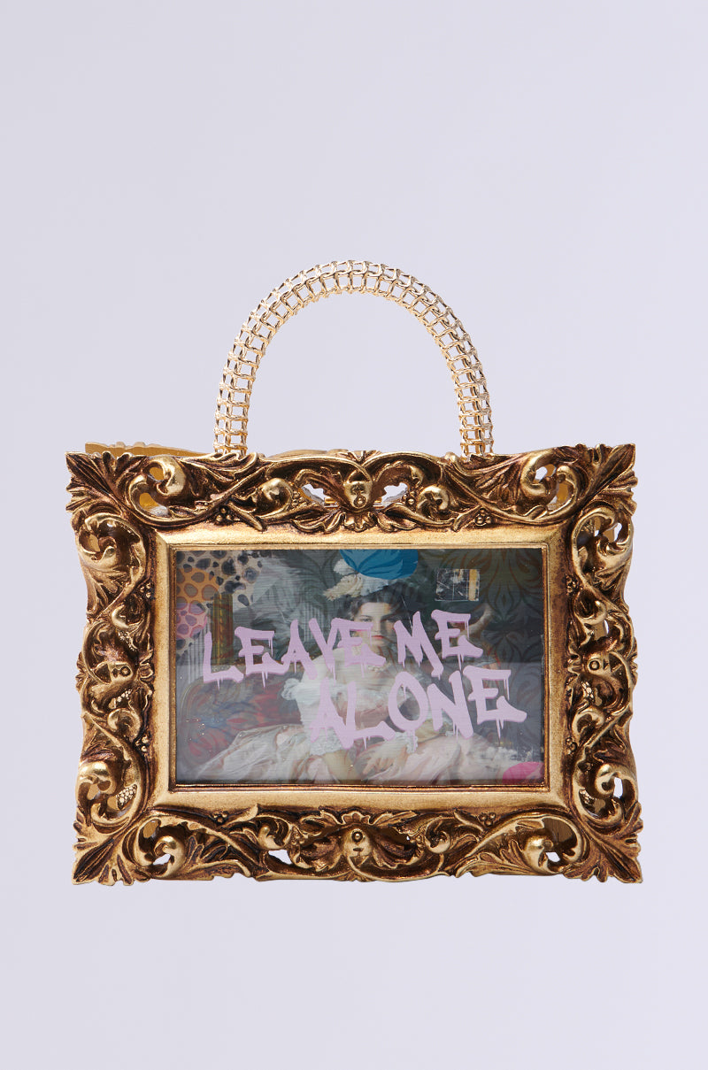 The FRAMED MIND PURSE is a novelty handbag with a baroque-style gold frame and chain handle, featuring vibrant artwork inside with "LEAVE ME ALONE" in bold pink graffiti over a detailed, colorful background.