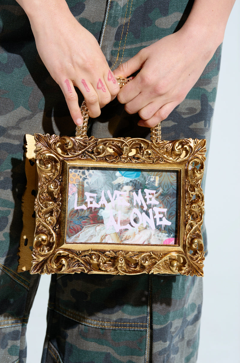 A person showcases the FRAMED MIND PURSE featuring a gold frame with a chain handle, adorned with "Leave Me Alone." They're dressed in camouflage pants, with "444" inked on their fingers. 1