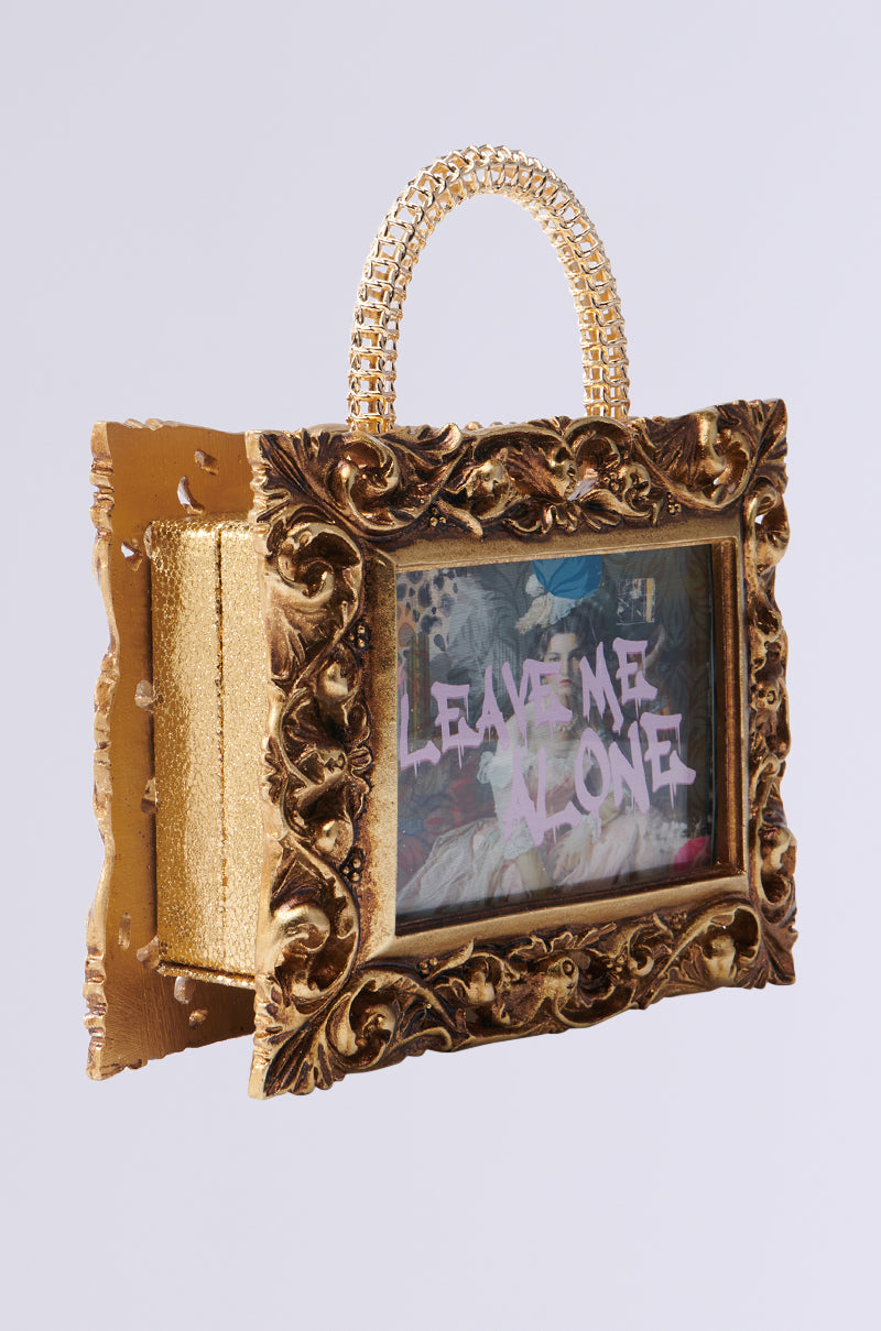 Introducing the FRAMED MIND PURSE: a gold ornate handbag with embossed floral details, featuring bold pink "LEAVE ME ALONE" text on a dreamy classical painting print. It resembles a picture frame and has an elegantly textured handle for a unique touch. 2
