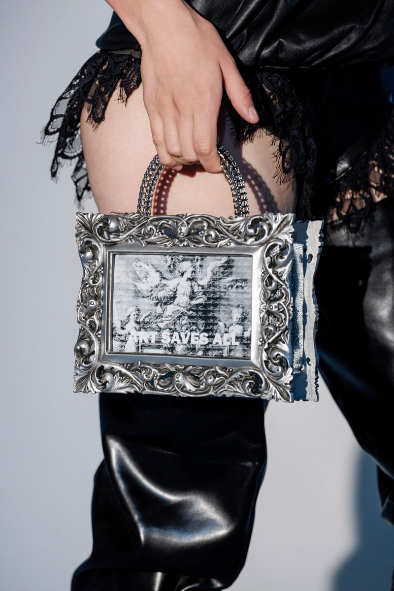 A person in black lace and leather holds the ART SAVES FRAME BAG—a striking silver baroque accessory featuring a black-and-white image with the words "ART SAVES" inside.