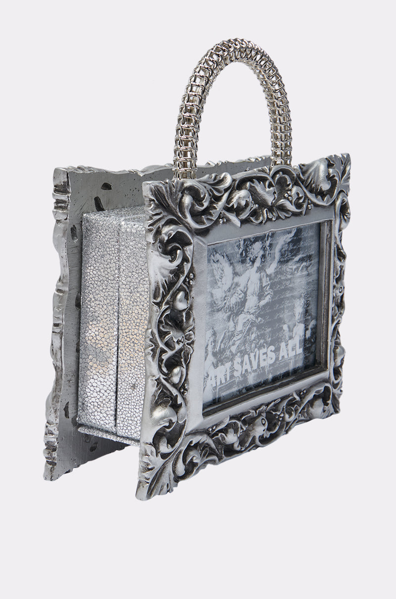 Make a statement with the ART SAVES FRAME BAG—shaped like an ornate silver picture frame with a textured handle, it features “ART SAVES ALL” over a black-and-white image and metallic textured side panels.
