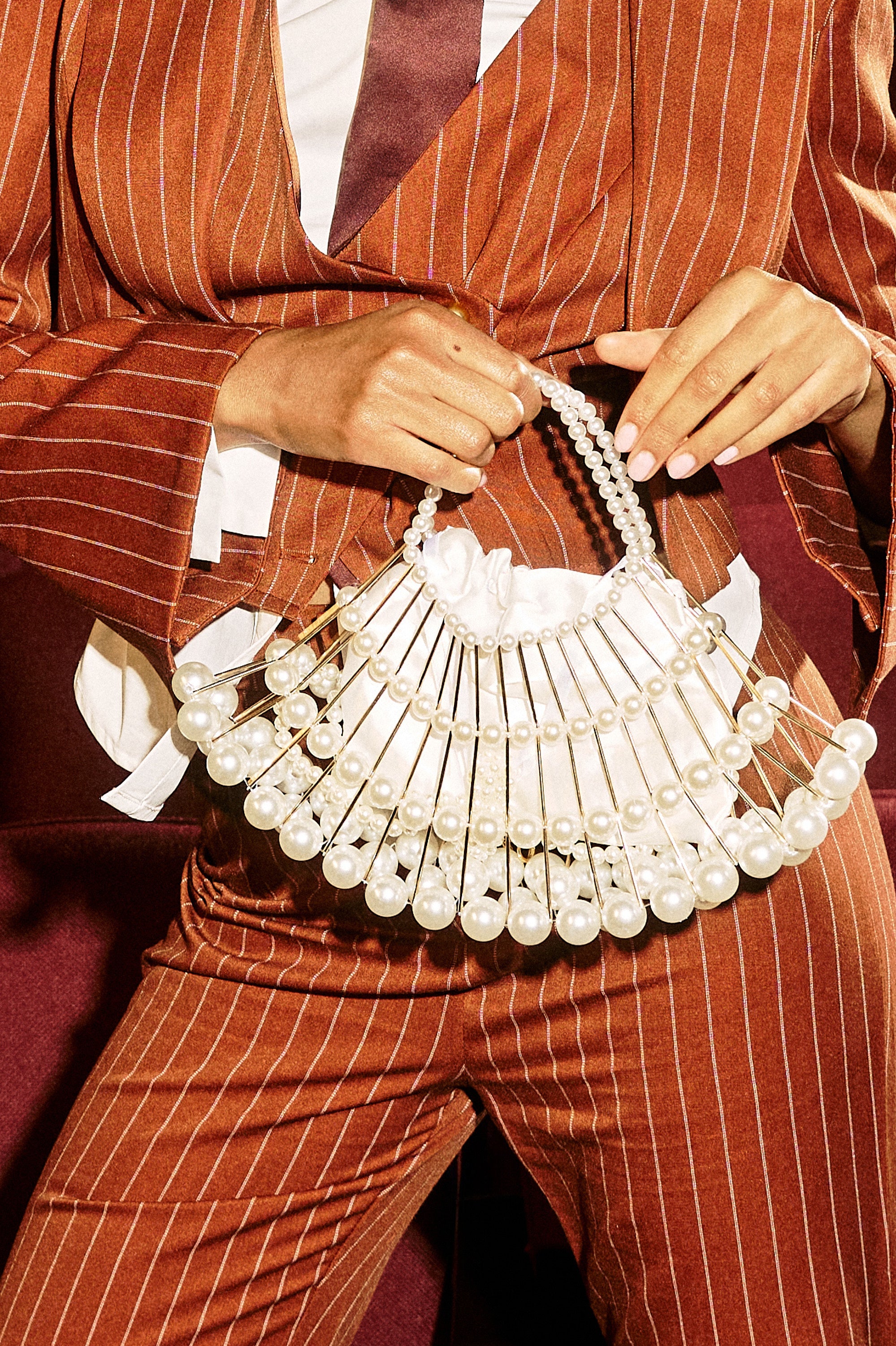 A person in a rust pinstripe suit holds the DIVINE DREAMS PEARL PURSE, featuring large beads and a white satin pouch, with their shirt partially untucked and a maroon tie visible. 4