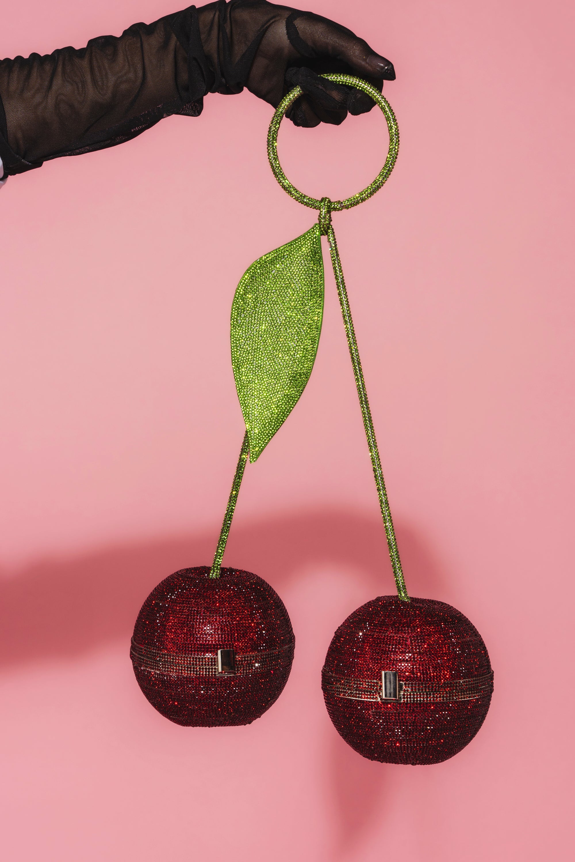 A gloved hand holds the POP MINE PURSE, a novelty handbag shaped like two red cherries with a green stem and leaf, set against a pink background. 0