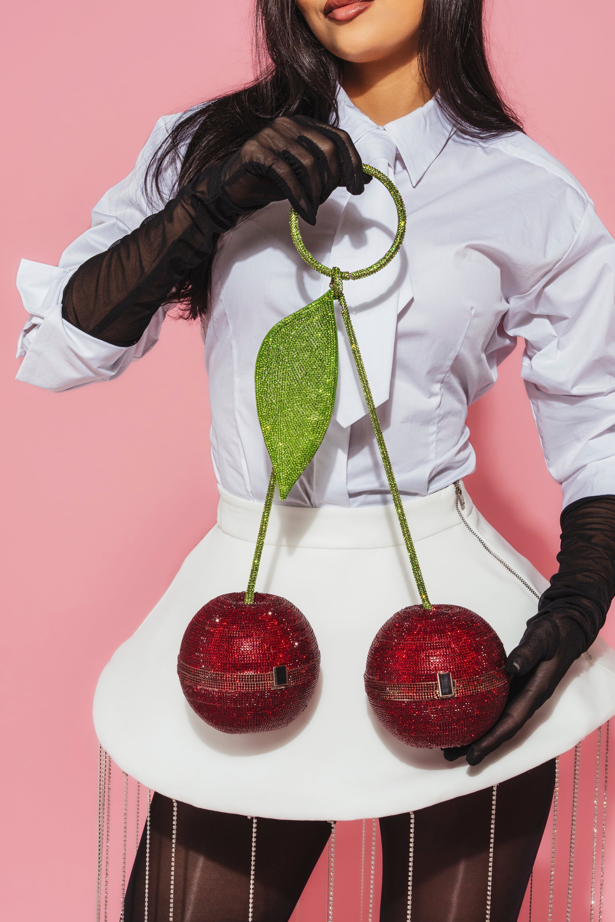 A woman in a white blouse, skirt, black gloves, and sheer tights holds the shiny POP MINE PURSE—a playful cherry-shaped novelty bag with a green leaf—against a pink background. Her face is partially out of frame. 1