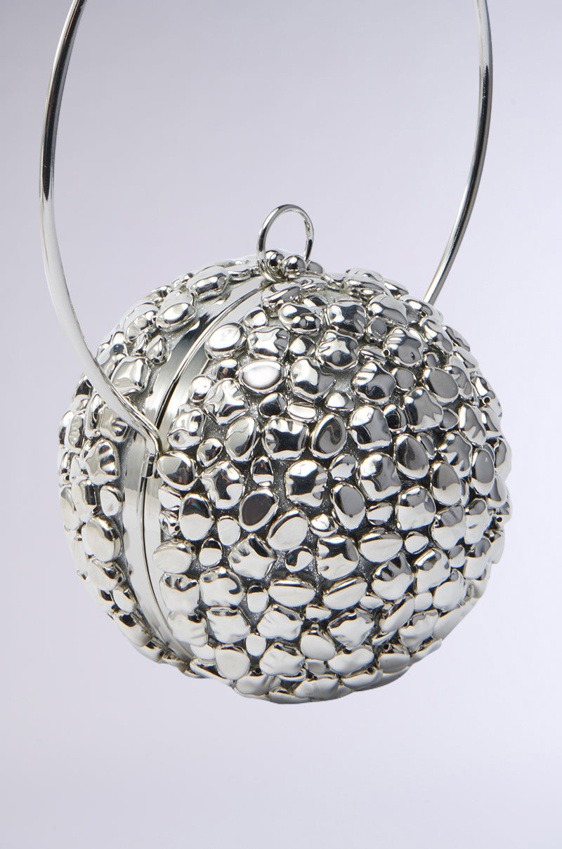 The TO THE FUTURE METAL PURSE is a statement accessory with a textured, pebble-like silver surface. It features a top clasp and circular handle, reflecting light softly against its neutral gray background, perfectly highlighting its intricate design.