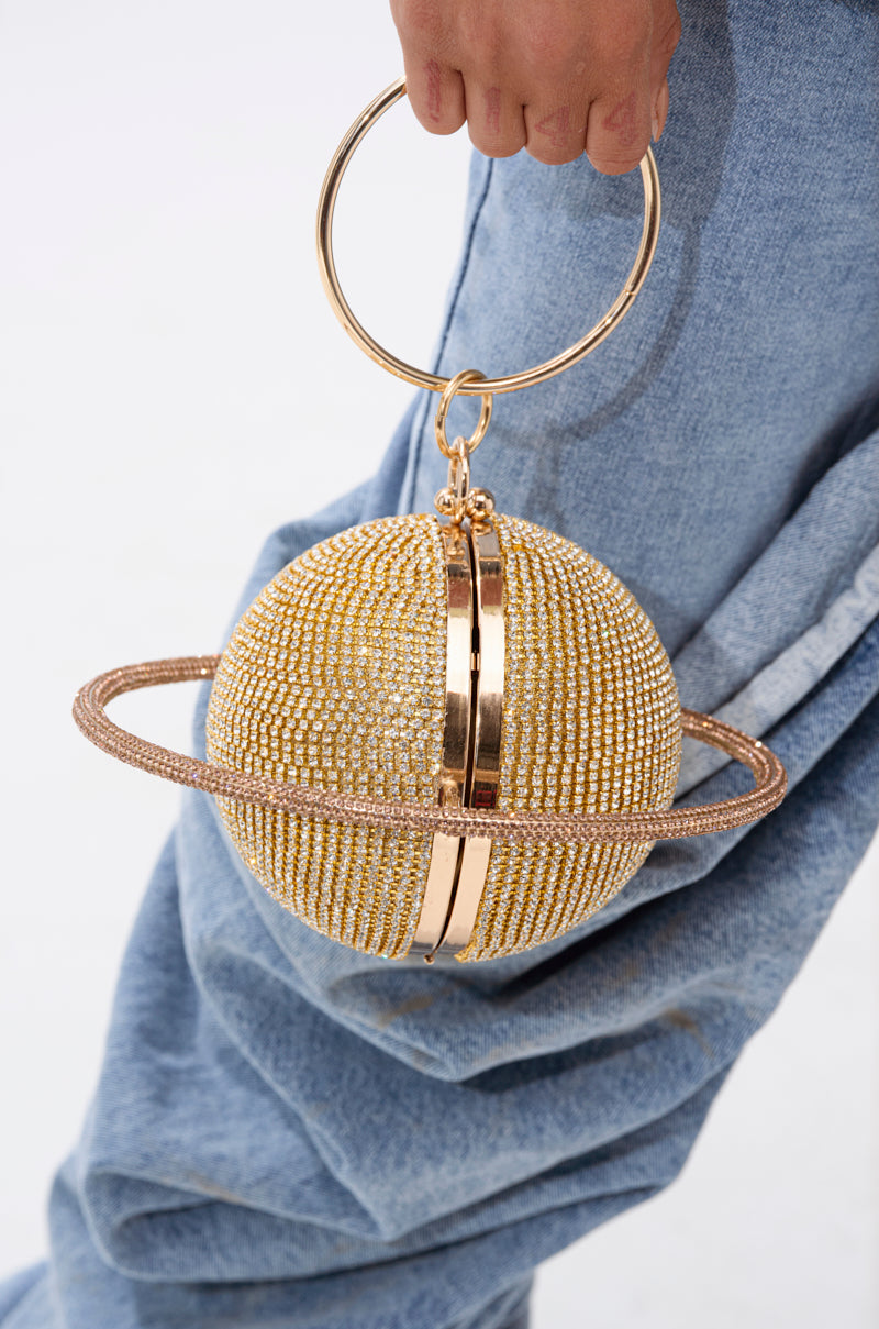 A person wearing blue jeans holds the OUTTA THIS WORLD PURSE, a Saturn-shaped accessory with sparkling rhinestones, a metallic ring handle, and circular bands resembling Saturn's rings. 1