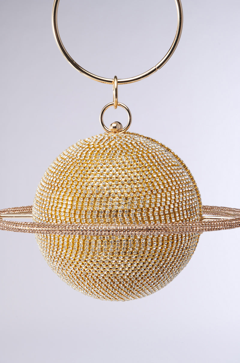 The OUTTA THIS WORLD PURSE is a gold metallic Saturn-shaped bag with dazzling rhinestones and a sparkling ring at its center, hanging from a gold hoop handle against a neutral background. 3