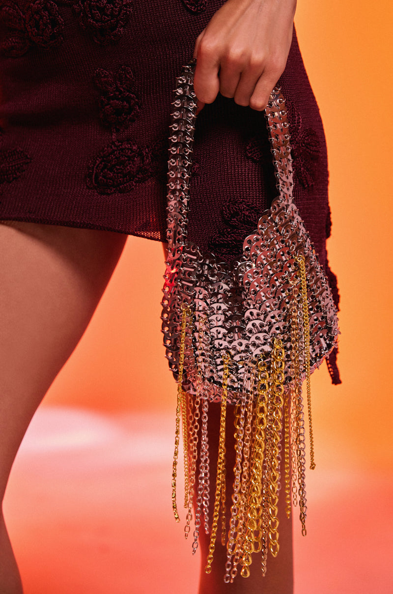 Against a warm orange background, a person holds the ABBY CHAIN PURSE—a compact silver handbag with dazzling mixed-metal chain fringe—while wearing a dark burgundy textured skirt.