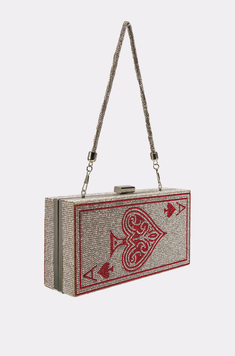 The PLAY YOUR CARDS CLUTCH is a rectangular silver rhinestone clutch with a beaded strap, showcasing a red and silver ace of hearts playing card design on the front. 3