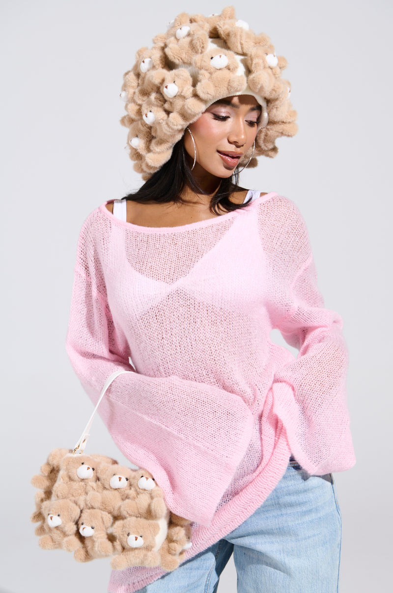 A woman in a pink knit sweater, faded blue jeans, and a beige hat adorned with plush bear appliques holds her BEAR WITH ME PURSE. The purse, featuring delightful plush bears, complements her quirky look against the gray backdrop.