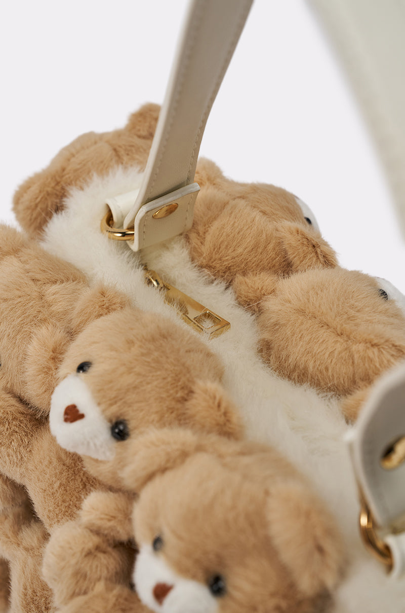A close-up of the BEAR WITH ME PURSE, a faux fur handbag featuring plush light brown and white teddy bears, gold hardware, and a stylish white strap. 3