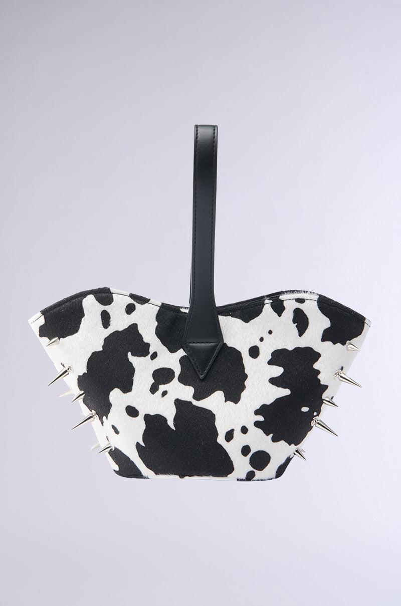 The BUT THIS AIN'T THE FARM MINI PURSE features a cow-print design, black leather handle, and gold spikes on the sides, making it a standout accessory displayed against a plain gray background.