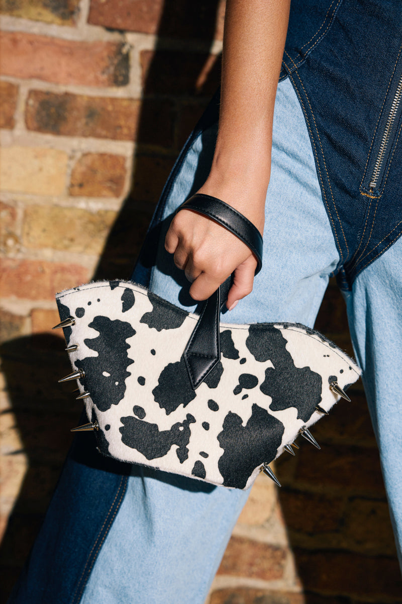 A person in blue denim holds the BUT THIS AIN'T THE FARM MINI PURSE—cow-print with black spikes and a black handle—in front of a brick wall, making a bold statement.