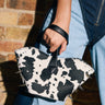 A person in blue denim holds the BUT THIS AIN'T THE FARM MINI PURSE—cow-print with black spikes and a black handle—in front of a brick wall, making a bold statement.